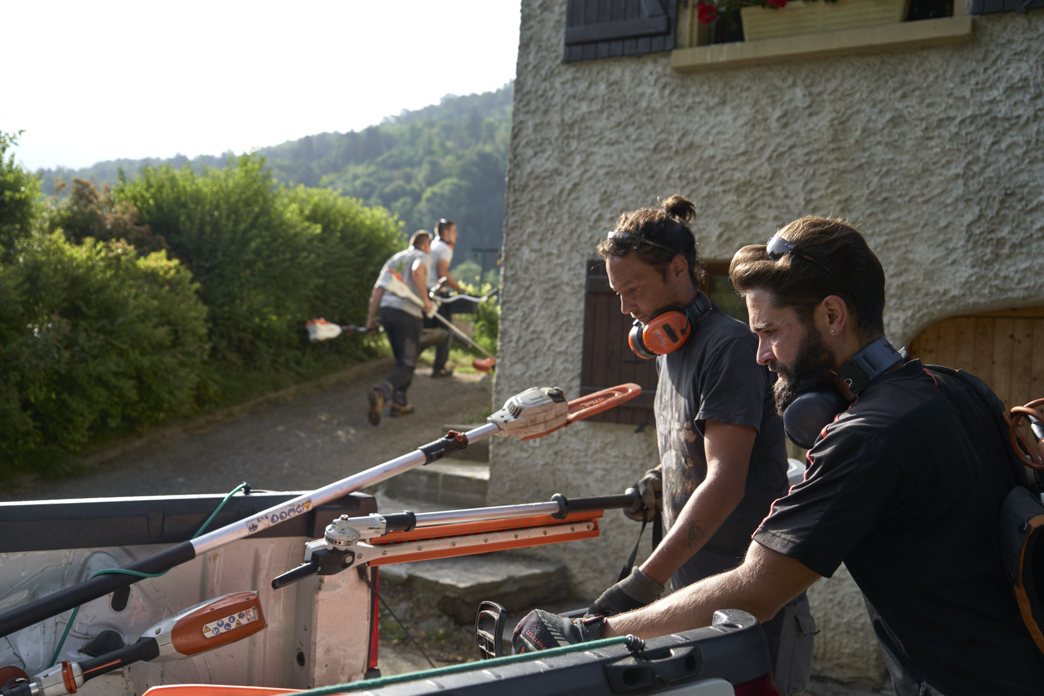 Dos jardineros paisajistas descargan las máquinas de batería STIHL de un coche; en segundo plano hay otros trabajadores 