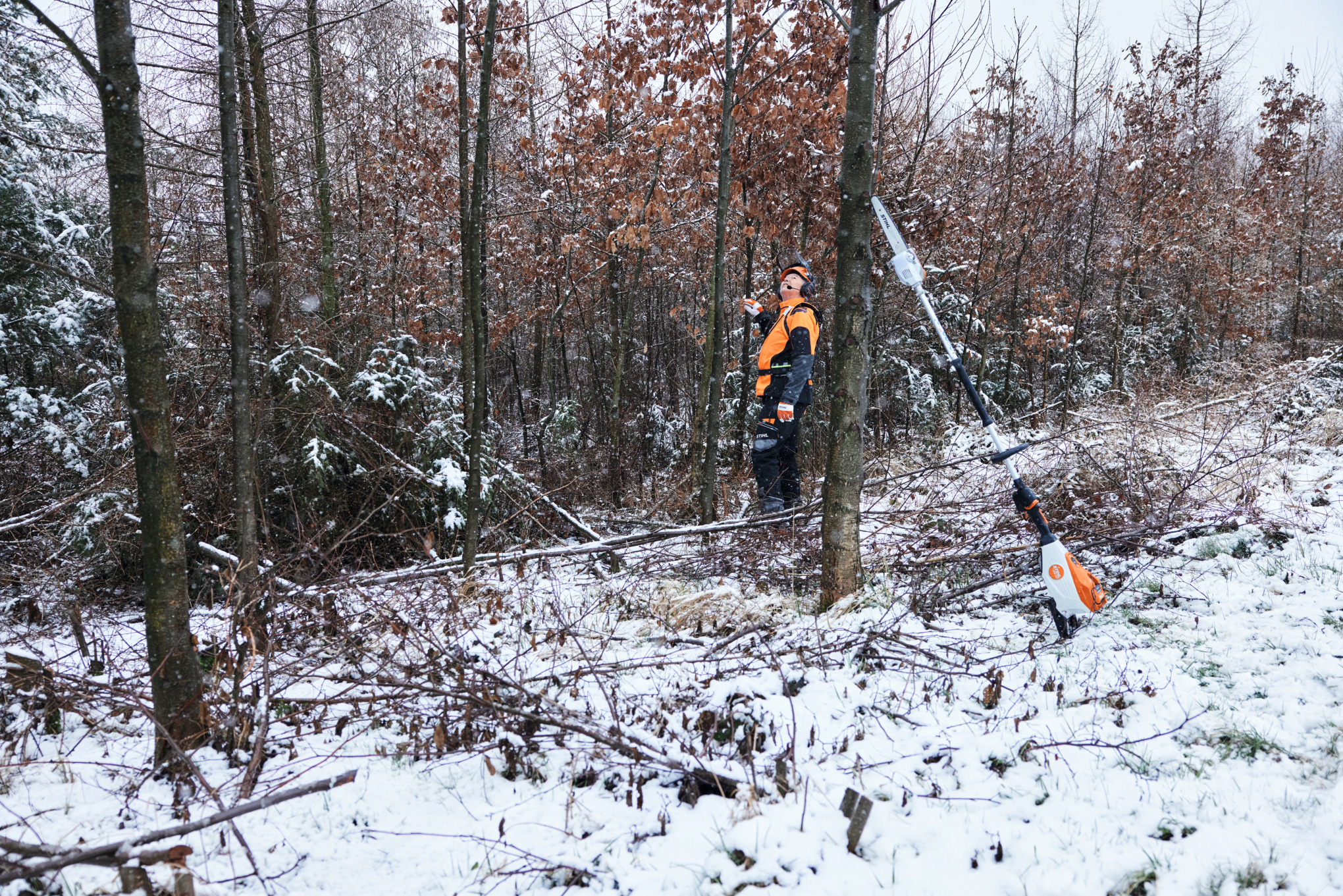 Un hombre trabaja con equipo de protección laboral en un bosque nevado; en primer plano hay una podadora de altura de batería STIHL HTA 150 apoyada sobre un tronco