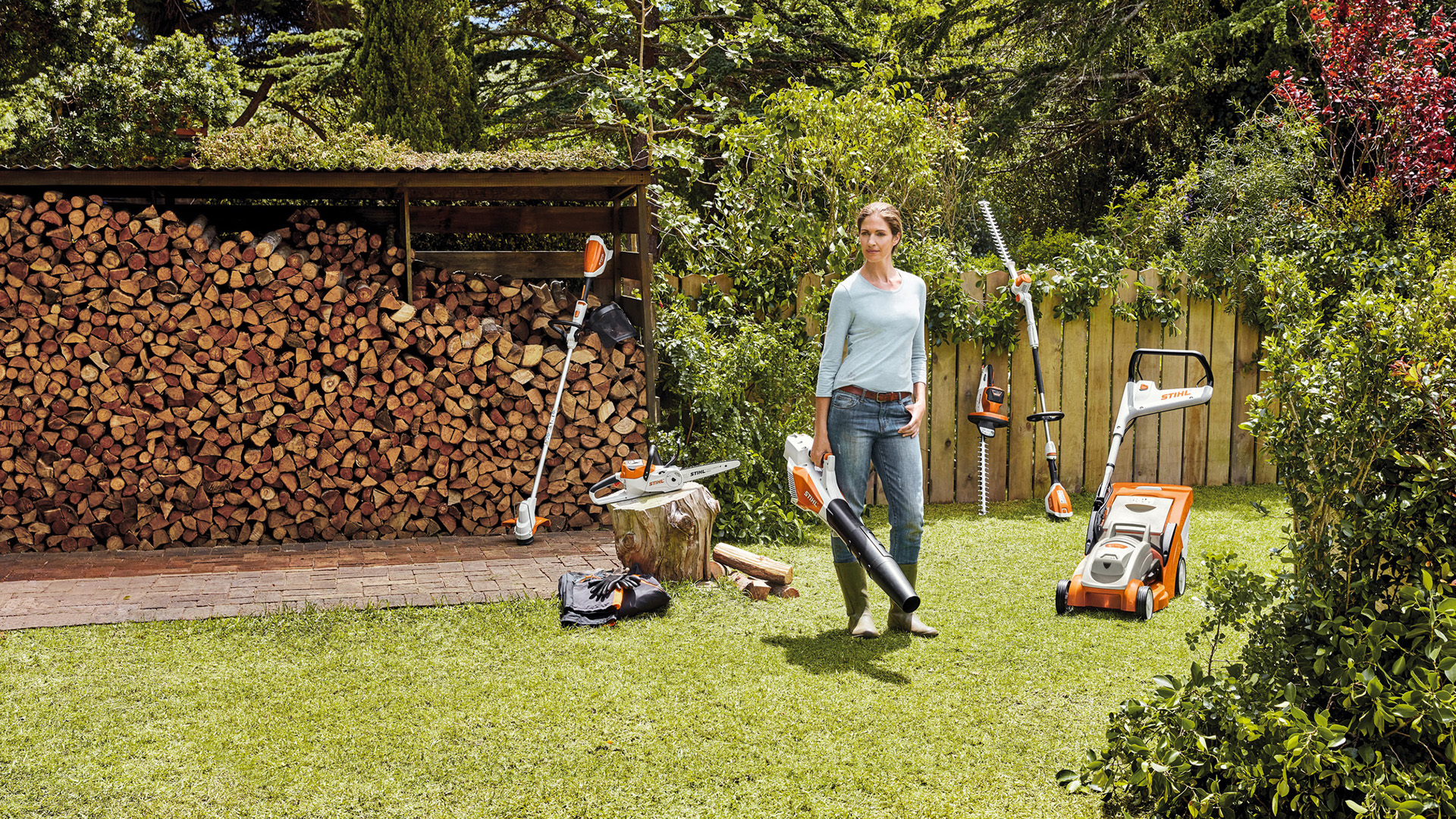 Una mujer de pie en un jardín con una selección de máquinas a batería del sistema STIHL AK